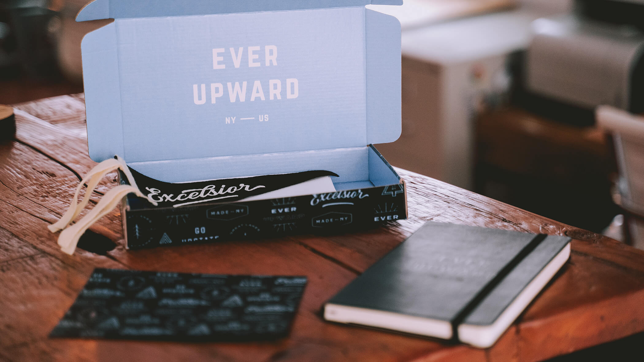 a box that says “Ever Upward” is open on a wooden table. Inside is a black pennant that reads “Excelsior”. On the table are two items: a black postcard with an Upstate NY graphic pattern, and a moleskine notebook that’s embossed to read “Ever Upward”.